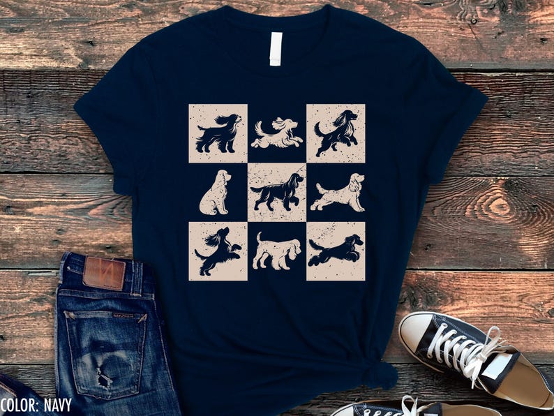 May include: Navy blue t-shirt featuring a grid of nine dog illustrations in various poses. The dogs are in a vintage style, with a distressed beige background. The shirt is paired with blue jeans and black sneakers.