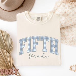 May include: An ivory-colored crewneck t-shirt featuring the text FIFTH in a blue and white gingham pattern with the word Grade written below in a dark gray script font. The shirt has a Comfort Colors brand label visible at the inner neckline.