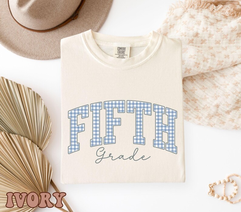 May include: An ivory-colored crewneck t-shirt featuring the text FIFTH in a blue and white gingham pattern with the word Grade written below in a dark gray script font. The shirt has a Comfort Colors brand label visible at the inner neckline.