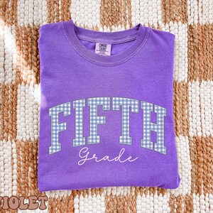 May include: A violet short-sleeved t-shirt featuring the text FIFTH in a blue and white gingham pattern, with the word Grade written below in white script. The shirt has a crew neckline and a Comfort Colors brand label on the inner collar.