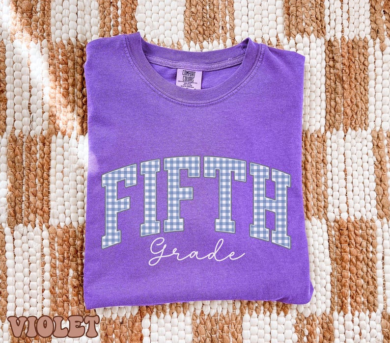 May include: A violet short-sleeved t-shirt featuring the text FIFTH in a blue and white gingham pattern, with the word Grade written below in white script. The shirt has a crew neckline and a Comfort Colors brand label on the inner collar.
