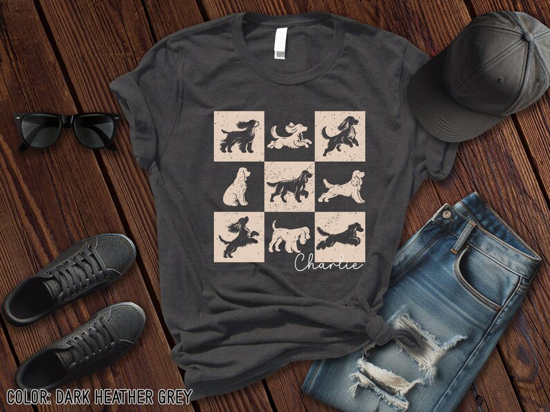 May include: Dark heather grey t-shirt featuring a grid of nine dog illustrations in various poses. The word "Charlie" is written in cursive below the grid. The shirt is paired with sunglasses, sneakers, a baseball cap, and jeans.