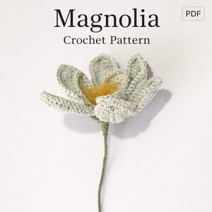 Pode incluir: Uma flor de magnólia de crochê, com pétalas cinza claro e um centro amarelo, presa a um caule verde. O texto "Magnolia Crochet Pattern" é exibido na parte superior da imagem. Artesanato feito à mão.