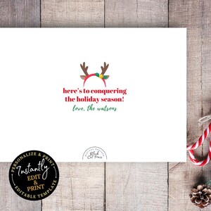 Op de afbeelding: Een witte wenskaart met de tekst "here's to conquering the holiday season!" in het rood en groen. De kaart heeft een rendiergeweimotief en wordt omringd door zuurstokken en dennennaalden.
