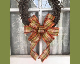 May include: A rustic grapevine wreath adorned with a large plaid bow. The bow features a mix of orange, green, and brown hues. The wreath is hanging on a white window with a sheer curtain backdrop.