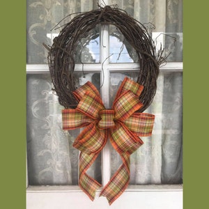 May include: A rustic grapevine wreath adorned with a large plaid bow. The bow features a mix of orange, green, and brown hues. The wreath is hanging on a white window with a sheer curtain backdrop.