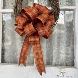 May include: A large decorative bow made of plaid ribbon in shades of orange, red, and green with metallic gold threading. The bow features multiple loops and two long, hanging tails, and is attached to a circular wreath made of brown twigs.