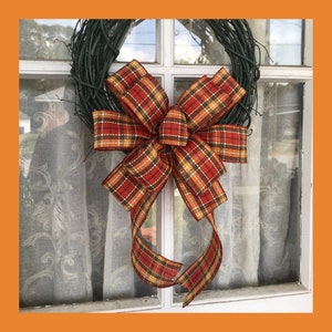 May include: A plaid ribbon bow in shades of orange, red, and yellow is tied to a grapevine wreath. The wreath is hanging on a window.