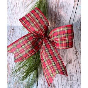 May include: A large, decorative bow made of red plaid fabric with green, yellow, and black accents. The bow is attached to a sprig of green pine needles, creating a festive holiday decoration. The bow's edges are finished with a red trim.
