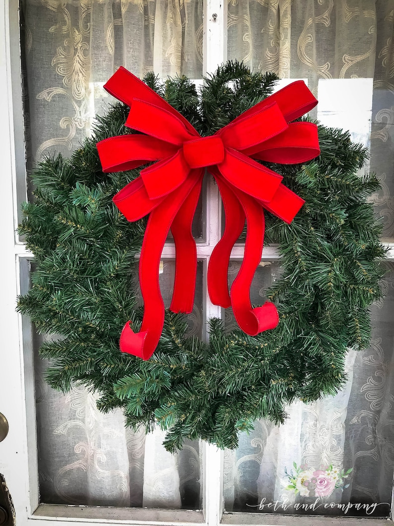 May include: A green Christmas wreath with a large red velvet bow. The wreath is hanging on a white door with a sheer curtain behind it.