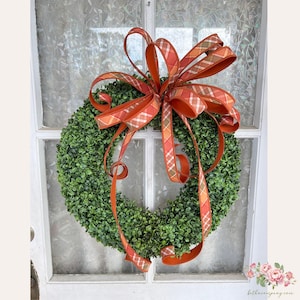 May include: A green boxwood wreath adorned with a large orange plaid ribbon. The wreath is hanging on a white-framed window with a frosted glass pane. The ribbon has a plaid pattern in shades of orange, brown, and cream.