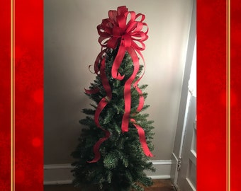 May include: A red ribbon Christmas tree topper with a large bow on top. The tree topper is on a small artificial Christmas tree.
