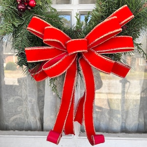 May include: A red velvet bow with gold trim is tied around a green evergreen wreath. The wreath is hanging on a white door.