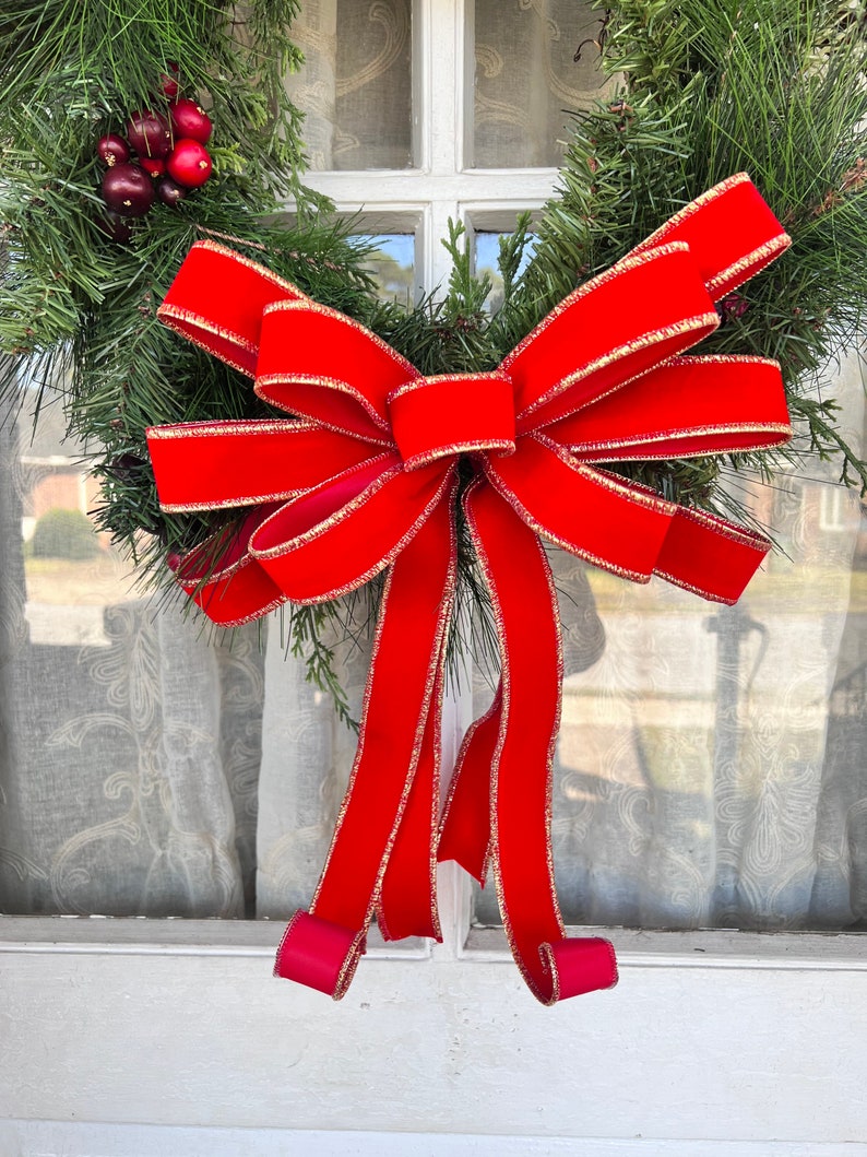 May include: A red velvet bow with gold trim is tied around a green evergreen wreath. The wreath is hanging on a white door.