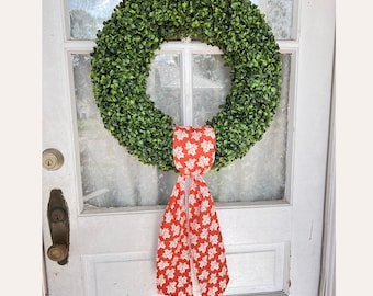 May include: A large, round green wreath with a dense, leafy texture hangs on a white door. A red ribbon with a white floral pattern is tied to the wreath, adding a pop of color. The door has a vintage-style handle.