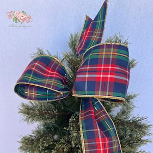 May include: A decorative bow made of fabric with a dark blue, green, red, and white plaid pattern. The edges of the ribbon are finished with a thin gold metallic trim. The bow is secured to the top of a green artificial pine tree branch.