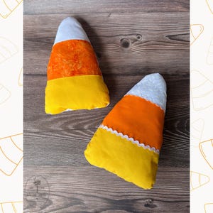 May include: Two plush candy corn decorations. Each candy corn has a white top, orange center, and yellow base. One candy corn has a white scalloped edge. The candy corn decorations are on a wooden surface.