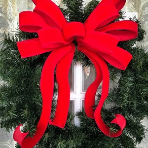 May include: A large, bright red velvet bow is tied around a green Christmas wreath. The bow is tied with a large, central knot and the tails of the bow are long and flowing.