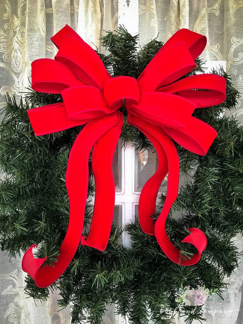 May include: A large, bright red velvet bow is tied around a green Christmas wreath. The bow is tied with a large, central knot and the tails of the bow are long and flowing.
