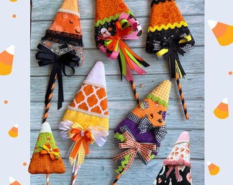 May include: Seven fabric candy corn decorations on sticks, each featuring unique patterns including orange, black, white, and purple prints. The ornaments are embellished with various ribbons, bows, lace trim, and rickrack, all attached to striped paper straws.