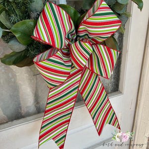 May include: A red, green, and white striped bow with a glittery finish. The bow is tied with a red ribbon and is hanging on a white door.