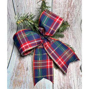 May include: A large, decorative plaid bow in red, navy blue, green, and gold. The bow is made of fabric with a gold trim and is attached to a small sprig of evergreen. The bow is suitable for holiday decorations.