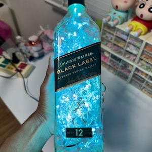 Puede incluir: Una botella de vidrio transparente con una etiqueta negra que dice "JOHNNIE WALKER. BLACK LABEL. BLENDED SCOTCH WHISKY John Walker & Sons AGED 12 YEARS" La botella está llena de luces blancas que emiten un brillo azul.