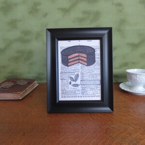 May include: A framed print of a chocolate cake with a dictionary page background. The cake is layered with chocolate and vanilla frosting. The print is in a black frame.