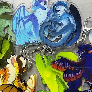 K&ouml;nnte beinhalten: Eine Sammlung farbenfroher Drachen-Acryl-Schl&uuml;sselanh&auml;nger. Die Schl&uuml;sselanh&auml;nger zeigen verschiedene Drachendesigns in Blau-, Gr&uuml;n-, Gelb- und Lilat&ouml;nen. Jeder Schl&uuml;sselanh&auml;nger hat einen silberfarbenen Verschluss zur Befestigung.