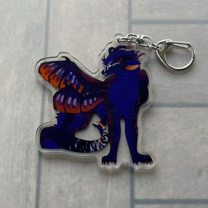 K&ouml;nnte beinhalten: Ein klarer Acryl-Schl&uuml;sselanh&auml;nger mit einem stilisierten Drachen mit blauen und orangefarbenen Fl&uuml;geln. Der Drache hat einen dunkelblauen K&ouml;rper mit orangefarbenen Akzenten und einen gestreiften Schwanz. Ein silberfarbener Schl&uuml;sselring und Verschluss sind befestigt.