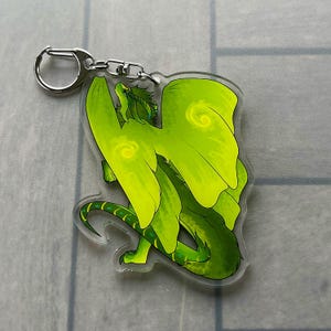 K&ouml;nnte beinhalten: Ein gr&uuml;ner Drachen-Schl&uuml;sselanh&auml;nger mit einem silbernen Schl&uuml;sselring. Der Drache hat gro&szlig;e Fl&uuml;gel und einen langen Schwanz mit einem Farbverlauf in Gr&uuml;nt&ouml;nen. Der Schl&uuml;sselanh&auml;nger ist aus klarem Acryl gefertigt.