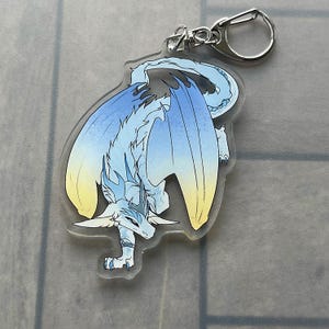 K&ouml;nnte beinhalten: Ein klarer Acryl-Schl&uuml;sselanh&auml;nger mit einer Drachenillustration. Der Drache hat blaue und gelbe Fl&uuml;gel, einen wei&szlig;en K&ouml;rper und eine silberfarbene Kette und einen Verschluss. Der Drache befindet sich in einer dynamischen Pose mit ausgebreiteten Fl&uuml;geln.