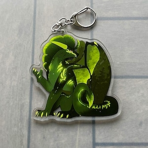 K&ouml;nnte beinhalten: Ein klarer Acryl-Schl&uuml;sselanh&auml;nger mit einer gr&uuml;nen Drachenillustration. Der Drache hat ein detailliertes Design mit Gr&uuml;nt&ouml;nen und Gelb. Der Schl&uuml;sselanh&auml;nger enth&auml;lt einen silberfarbenen Metallverschluss zum Befestigen an Schl&uuml;sseln oder Taschen.