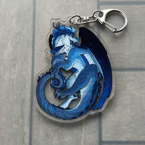 K&ouml;nnte beinhalten: Ein klarer Acryl-Schl&uuml;sselanh&auml;nger mit einer blauen Drachenillustration. Der Drache hat blaue und wei&szlig;e Schuppen, dunkelblaue Fl&uuml;gel und einen silberfarbenen Verschluss. Der Drache l&auml;chelt und hat einen langen Schwanz.