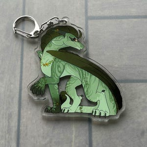 K&ouml;nnte beinhalten: Ein klarer Acryl-Schl&uuml;sselanh&auml;nger mit einem gr&uuml;nen Drachen mit dunkelgr&uuml;nen Fl&uuml;geln und Akzenten. Der Drache sitzt, mit einem silbernen Schl&uuml;sselanh&auml;ngerring oben befestigt. Der Drache hat eine Blumengirlande um den Hals.