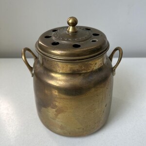 May include: A cylindrical brass container with a rounded base and two small side handles. The lid features a central knob and a series of circular ventilation holes arranged in a ring around the top, showing a weathered, metallic finish.