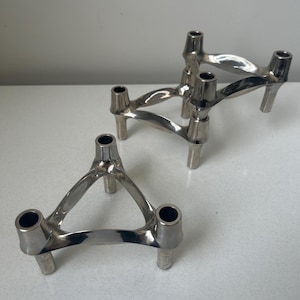 May include: Three silver-colored candle holders. Each holder has a triangular design with three cylindrical openings for candles and three short legs. The holders are made of a shiny, metallic material and are arranged on a white surface.