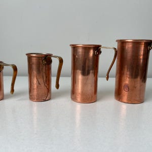 May include: Four copper measuring cups of varying sizes with brass handles. The cups are arranged in a row, showcasing their graduated sizes. The copper has a warm, reddish-brown hue, and the handles are curved.