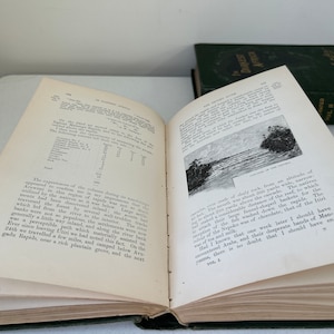 May include: An open book with aged, yellowed pages, featuring printed text and a black and white illustration of a river scene. A second, closed book with a dark green cover is visible in the background. The books appear to be antique.