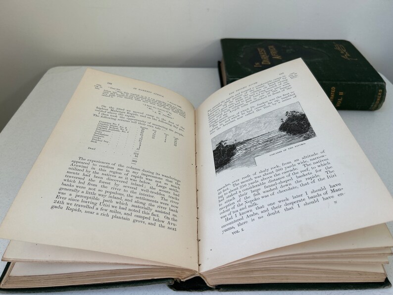 May include: An open book with aged, yellowed pages, featuring printed text and a black and white illustration of a river scene. A second, closed book with a dark green cover is visible in the background. The books appear to be antique.