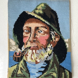 May include: Needlepoint portrait of a man with a pipe, wearing a green and black hat and jacket. The man has a white beard and pink cheeks. The background is a light blue color, and the piece is framed with a tan border.