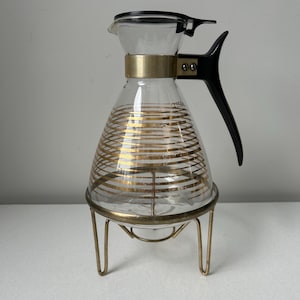 May include: A clear glass carafe featuring horizontal gold stripes, a black plastic handle, and a black lid. The carafe is supported by a gold-toned metal stand with three legs. The glass body has a tapered neck and a wider base for stability.