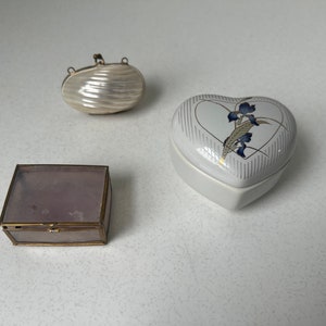 May include: Three decorative boxes: a small, pearlescent, oval-shaped box, a heart-shaped white box with a floral design, and a rectangular, lavender-coloured box with a gold frame. These boxes are ideal for storing small items.