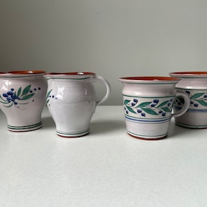 May include: Four ceramic items: two mugs and two jugs. The mugs and jugs are white with a pinkish tint, adorned with blue berries and green leaves. Each has a brown rim and a green stripe near the base.