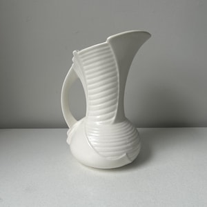 May include: A white ceramic pitcher with a curved handle and a flared spout. The pitcher has a textured, ribbed design on the body and a smooth, glossy finish. The pitcher is likely used for serving beverages or as a decorative item.