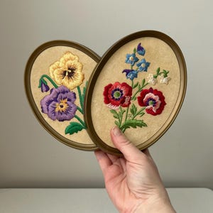 May include: Two oval framed floral embroidery pieces. One features a yellow and purple pansy, the other red and blue flowers. The frames are gold-toned, and the background is a beige fabric.