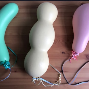 May include: Three painted wooden balloon-shaped sculptures in pastel colors: mint green, cream, and pink. Each has a ribbon tied at the base. The sculptures are displayed on a wooden surface.
