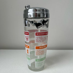 May include: A clear glass cocktail shaker with a metallic silver lid. The exterior features black graphic illustrations of barware and red, orange, and green text blocks detailing recipes for drinks like Whiskey Sour, Martini, and Gin & Tonic.