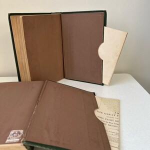 May include: Open antique book with a green spine and brown interior covers. A cream-colored paper is partially inserted into a pocket inside the book. The book's pages are aged and yellowed.