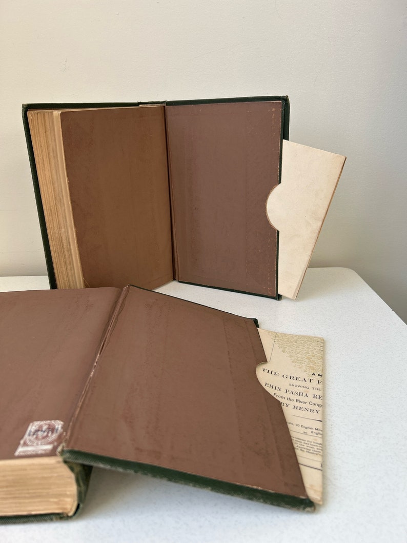 May include: Open antique book with a green spine and brown interior covers. A cream-colored paper is partially inserted into a pocket inside the book. The book's pages are aged and yellowed.